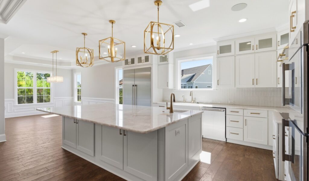 Spacious kitchen in custom home, featuring white cabinetry, marble island, gold light fixtures, and hardwood floors, with abundant natural light.