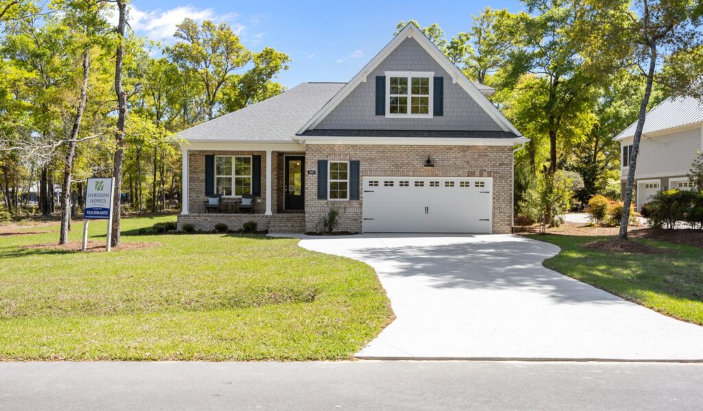 Brick custom home with a two-car garage, porch, and front lawn, surrounded by trees under a clear blue sky; Horizon Homes sign in yard.