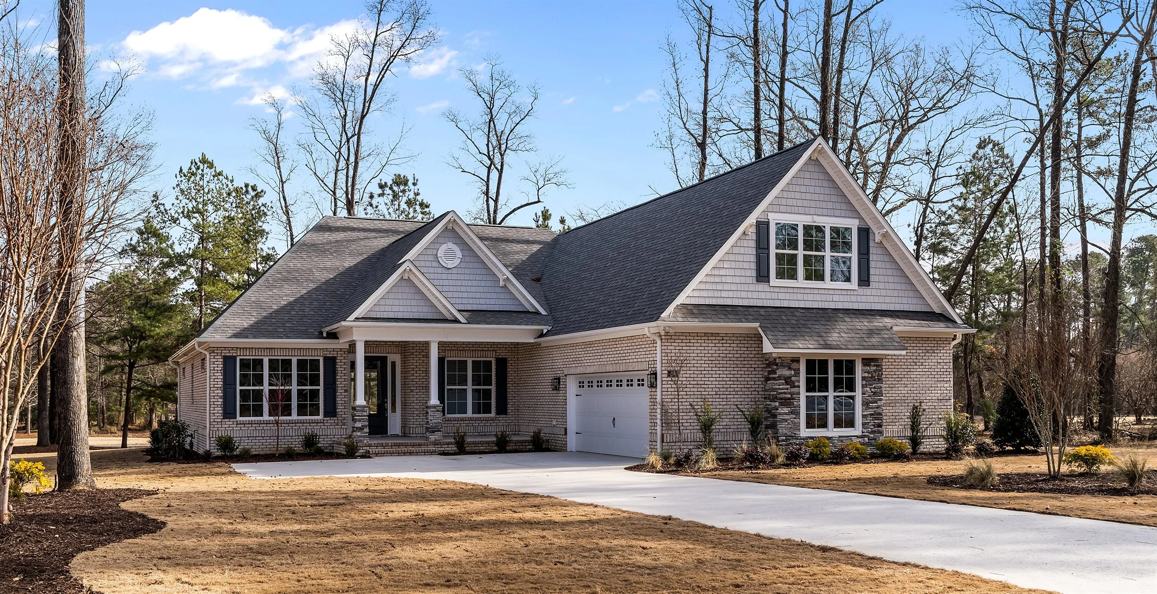 Single-story brick home with gabled roof, large windows, and a two-car garage, set in a wooded area. Ideal design for a home builder.