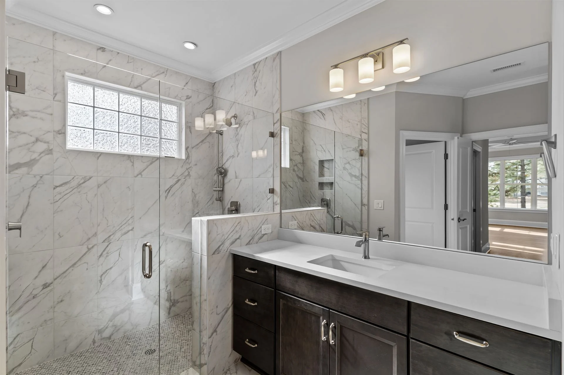 Modern bathroom with marble shower, dark wood vanity, and glass partition, designed by a home builder. Natural light from frosted window.