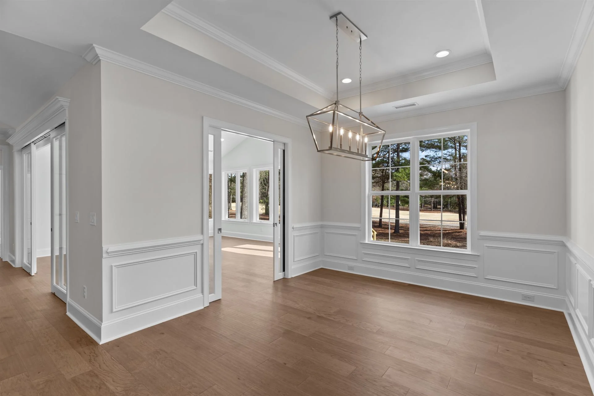Modern dining room with tray ceiling, large window, and chandelier, showcasing craftsmanship of a home builder. White walls, wooden floors.