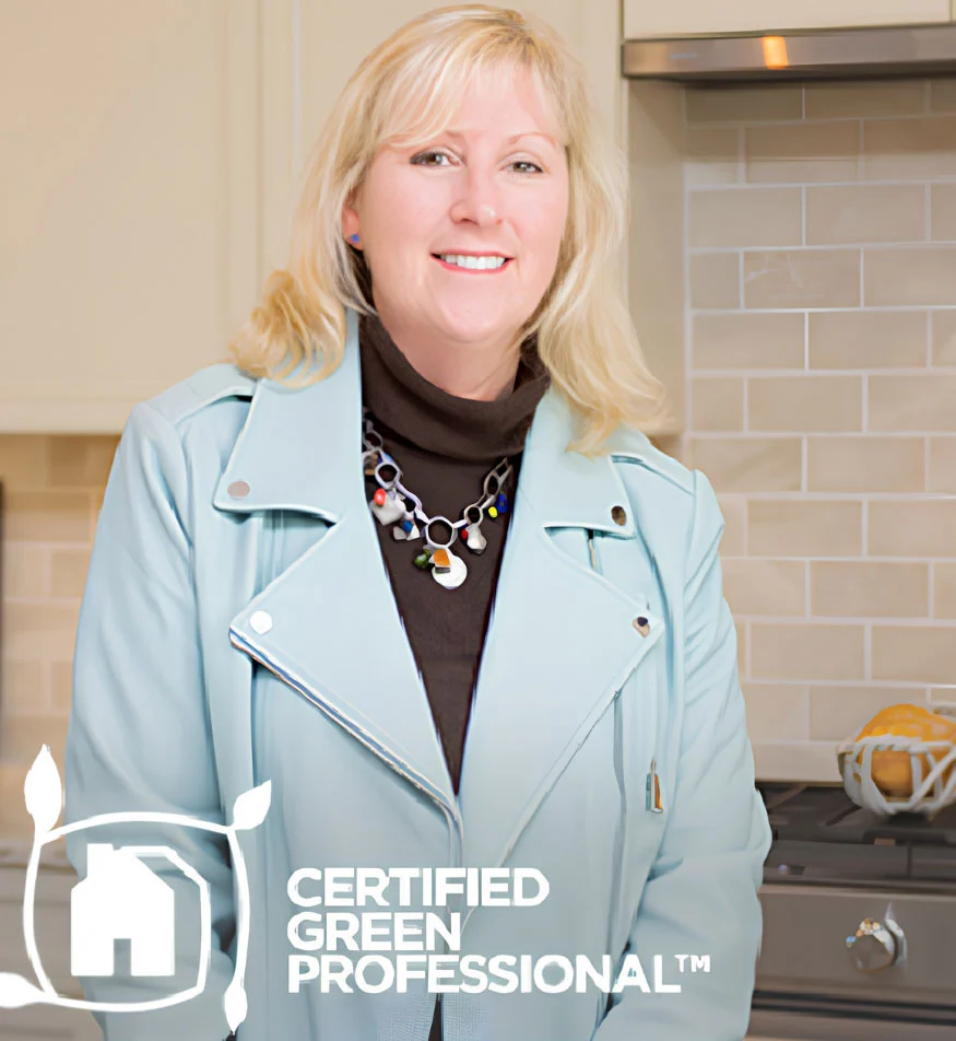 Assets_08 Smiling woman in a light blue jacket with "Certified Green Professional" logo, standing in a modern kitchen, representing eco-friendly home builder.
