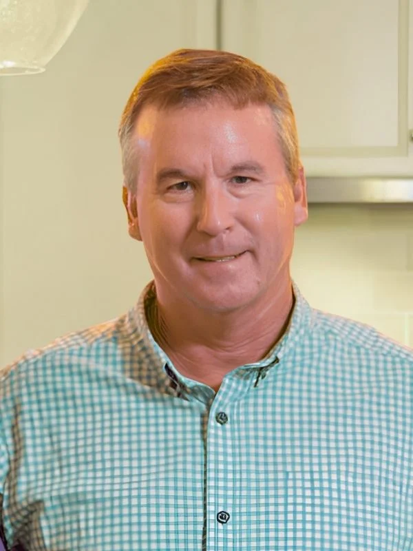 Smiling man in a checkered shirt stands in a kitchen, possibly a home builder.