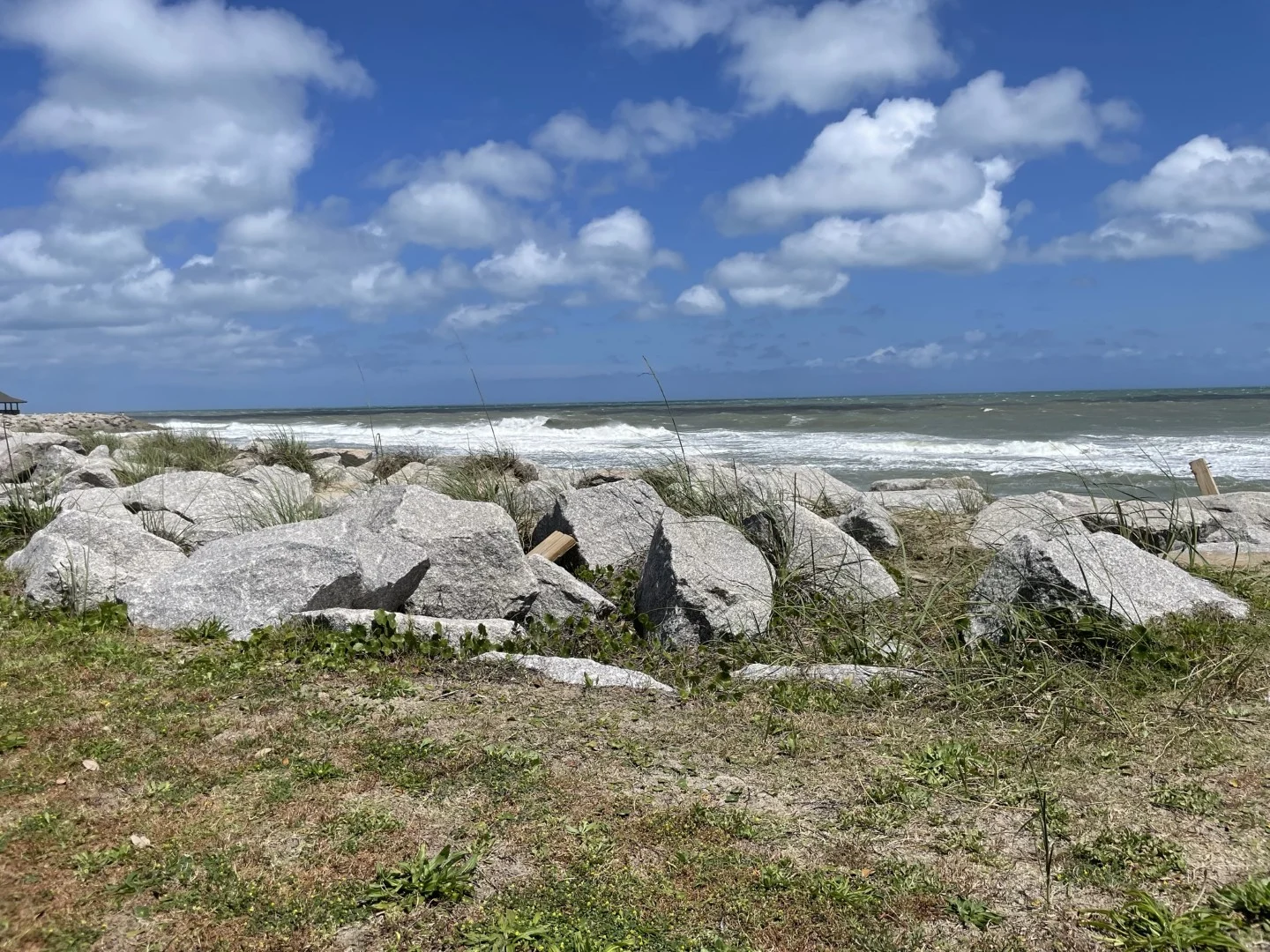 Coastal landscape with large rocks, grassy foreground, and waves beneath a blue sky with clouds. Ideal site for a retreat by a home builder.