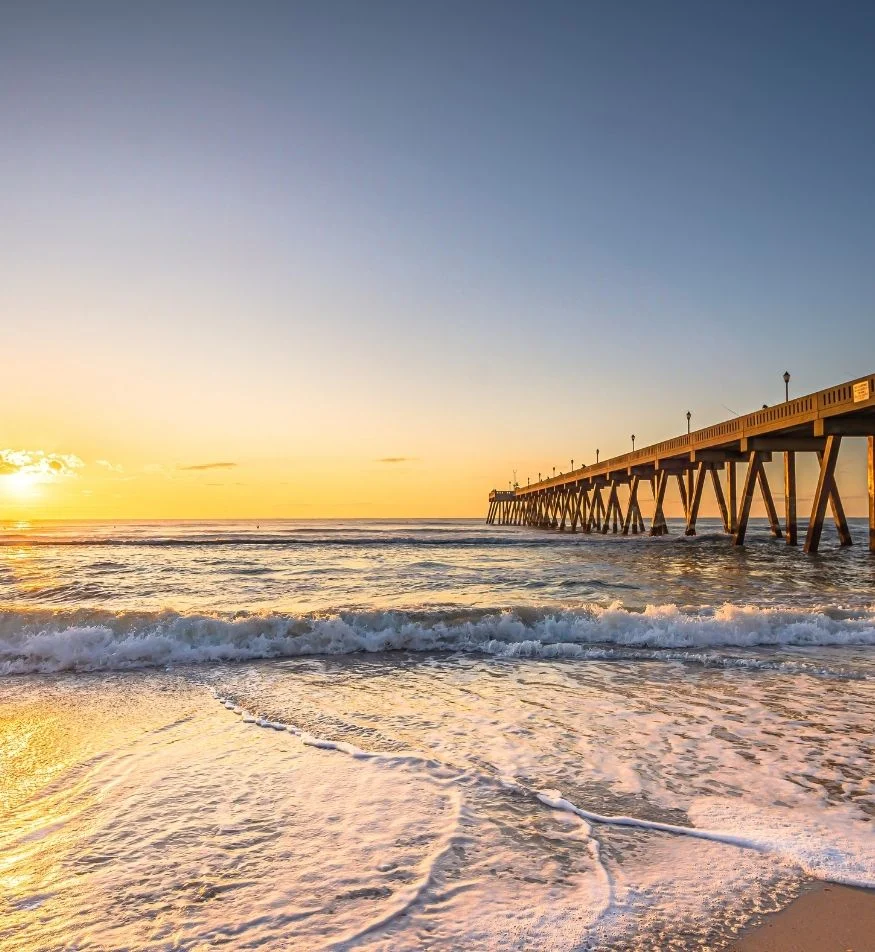 Sunset over ocean waves near a long pier, offering serene inspiration for a home builder looking to create coastal retreats.