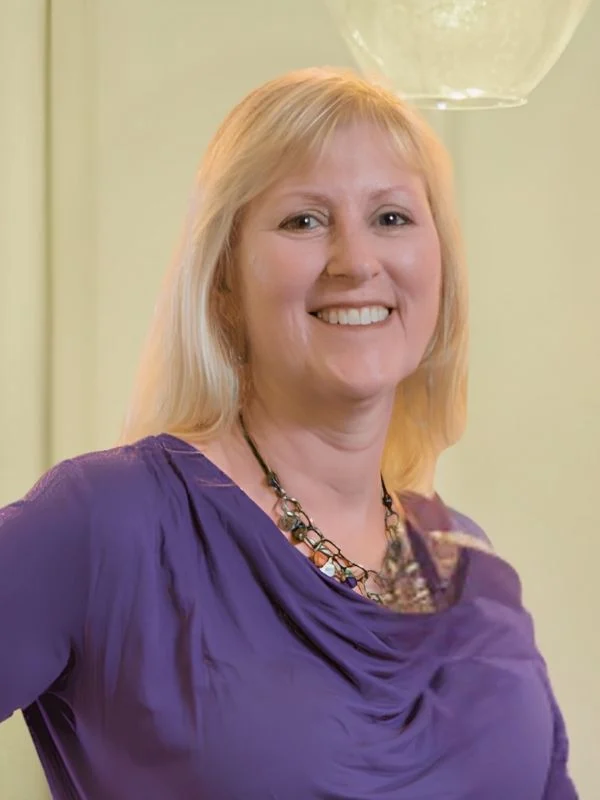 Smiling woman with blonde hair and purple top, wearing a beaded necklace. Bright background, warm ambiance.