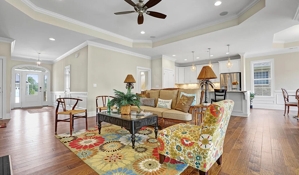 Spacious living room with colorful rug, cozy seating, and modern kitchen, showcasing home builder craftsmanship. Ceiling fan above.