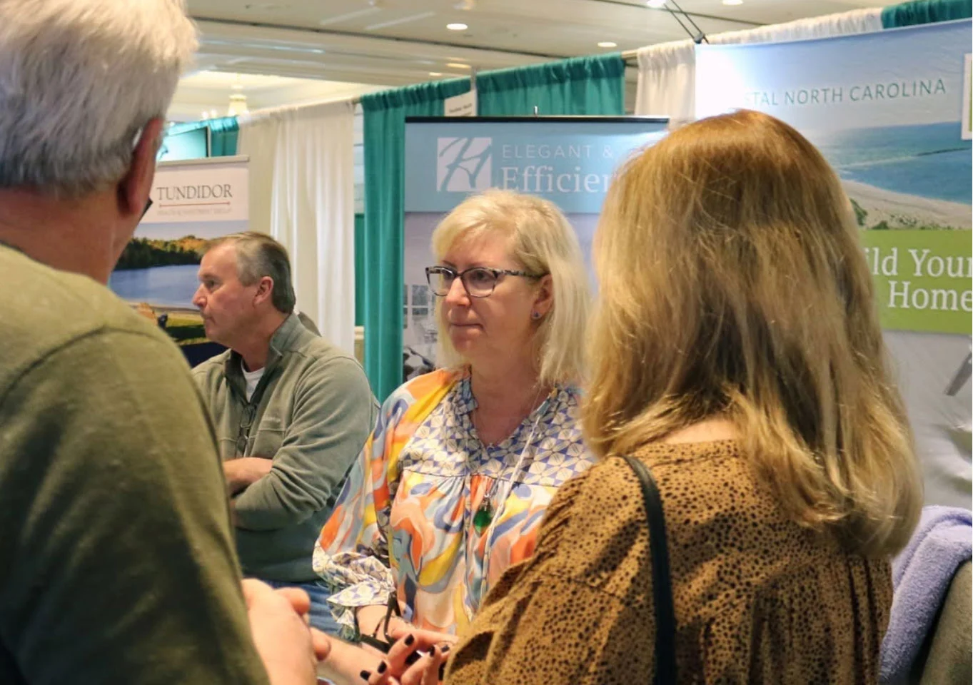 Woman in patterned blouse discusses with visitors at a home builder expo, promotional banners visible in the background.