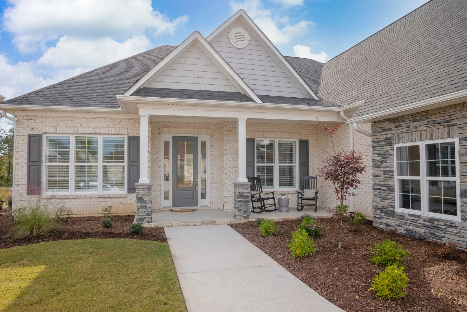 gallery-exteriors (29) Modern home exterior with gabled roof, brick facade, and covered porch; landscaping includes shrubs and young trees. Ideal for a home builder.