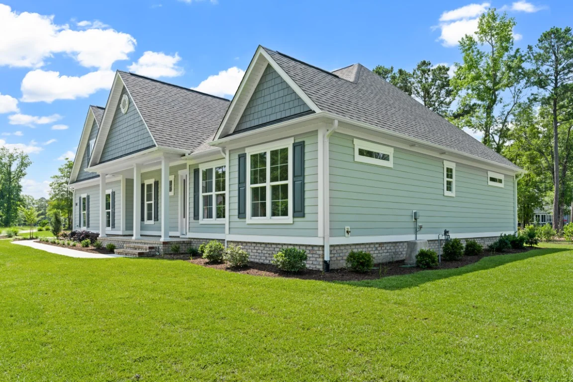 gallery-exteriors (32) Modern single-story house with light blue siding, gabled roof, lush lawn, surrounded by trees; work of a skilled home builder.