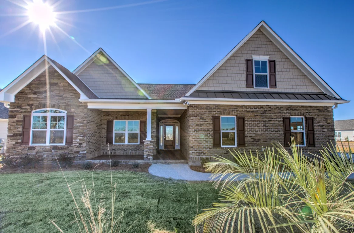 gallery-exteriors (6) Single-story brick home with brown shutters, gabled roof, and front porch in a sunlit yard, showcasing home builder craftsmanship.