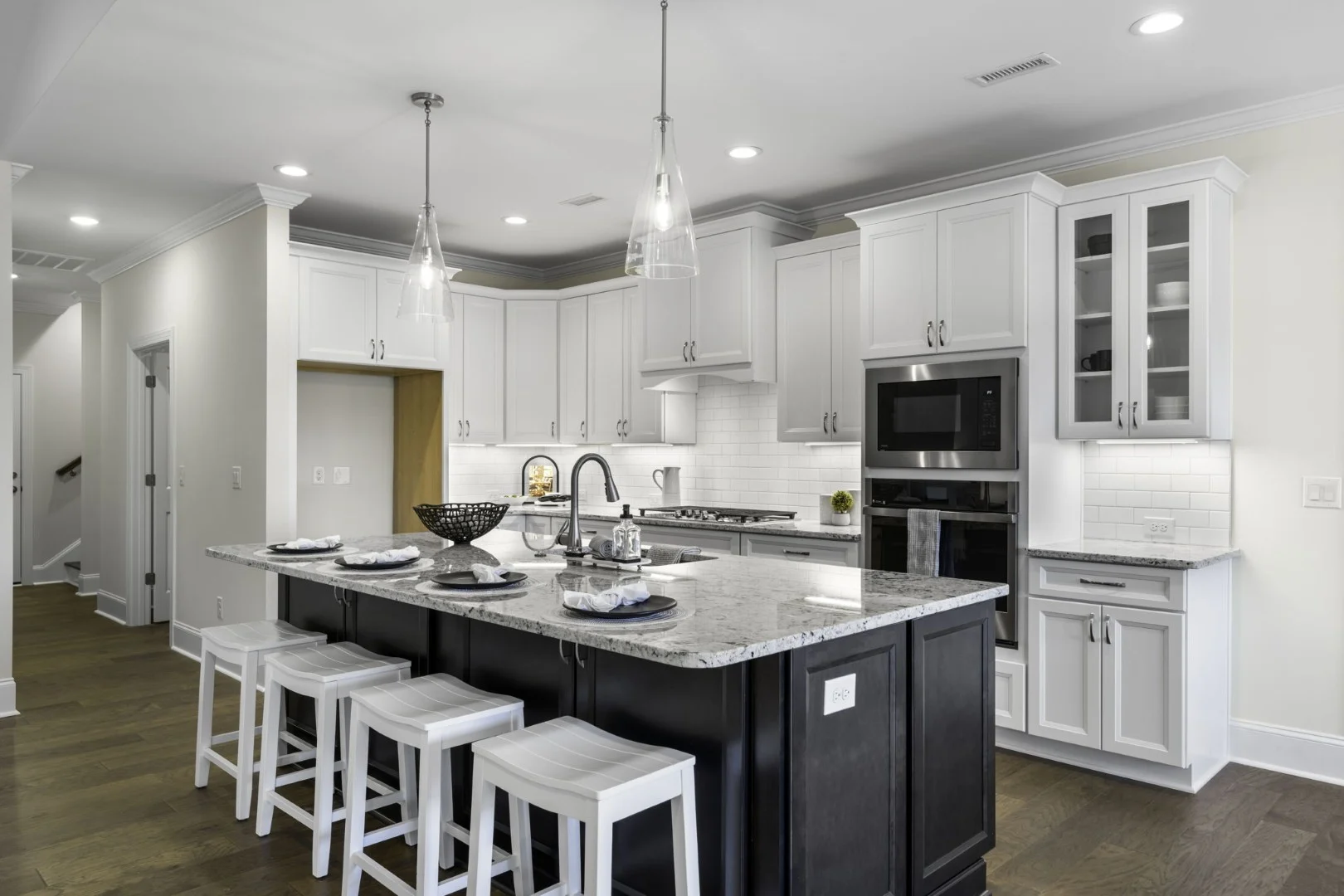 Modern kitchen with granite island, white cabinets, and stainless appliances. Pendant lights and white stools enhance this home builder's design.