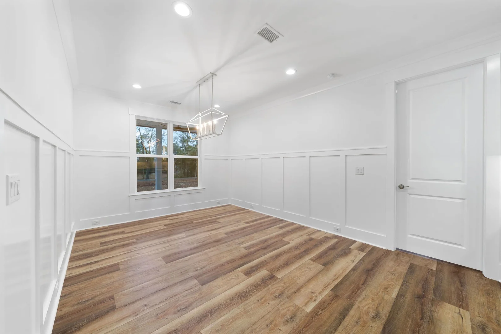 gallery-kitchen (12) Bright, empty room with wood flooring, white wainscoting, modern chandelier, and large window, showcasing home builder's design skills.