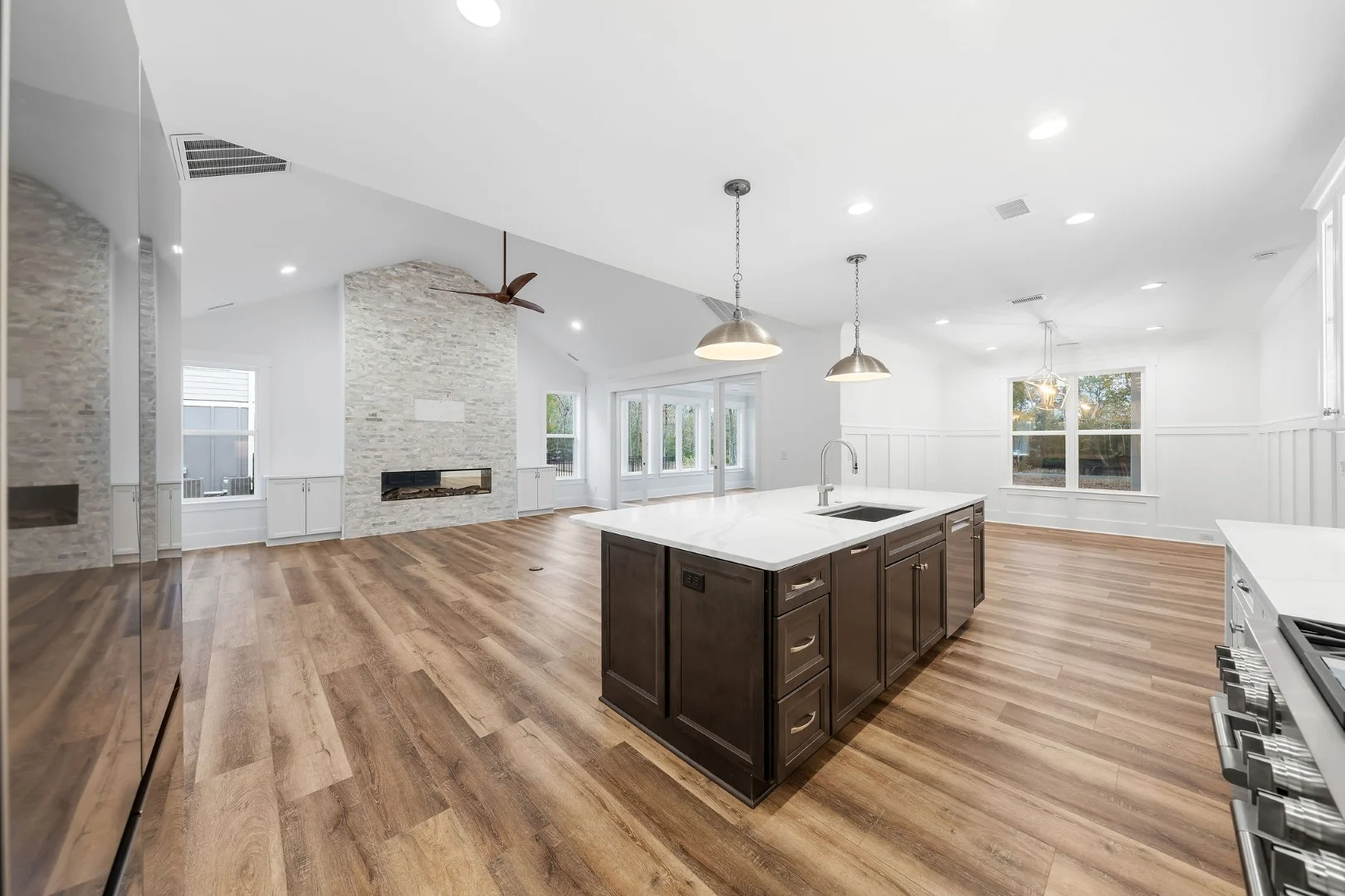 gallery-kitchen (17) Spacious modern kitchen with island, pendant lights, and wooden floors by home builder, featuring a stone accent wall with fireplace.
