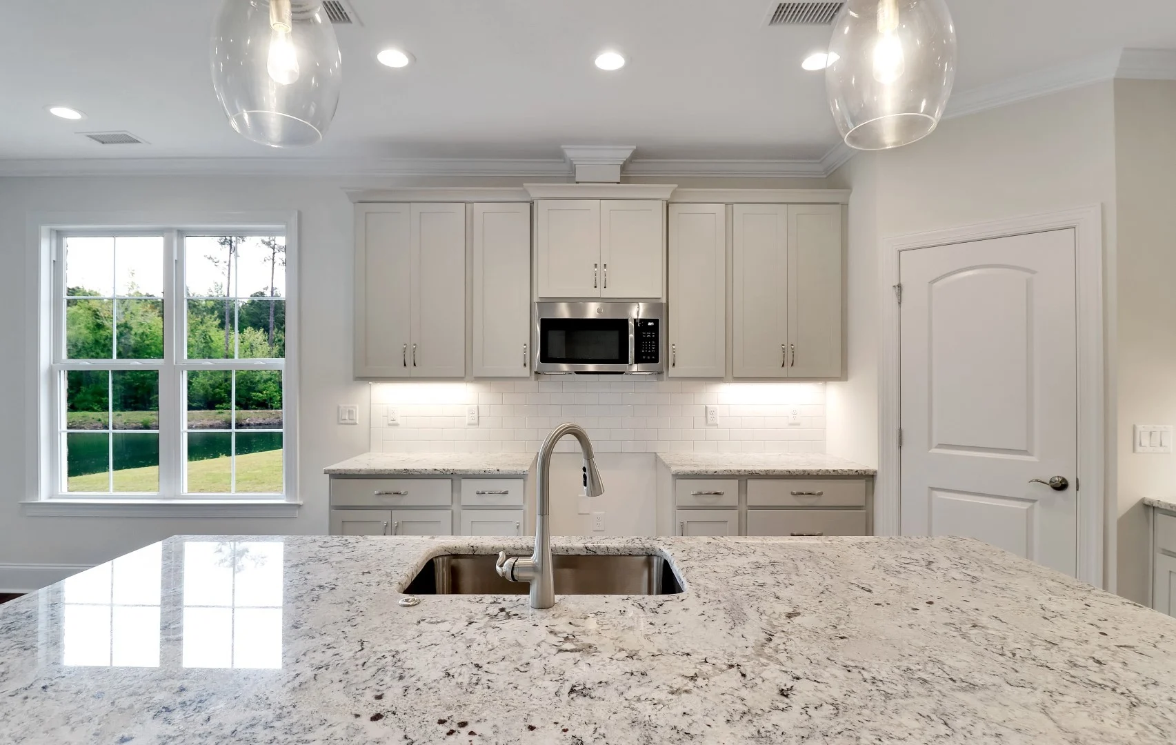 gallery-kitchen (30) Modern kitchen with marble island, stainless steel sink, and white cabinets. Large window offers natural light, designed by home builder.
