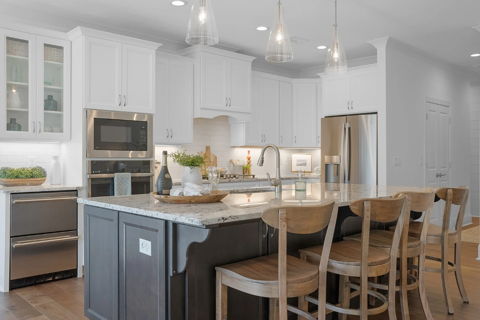 gallery-kitchen (32) Modern kitchen with white cabinets, granite island, wooden chairs, pendant lights, stainless steel appliances; ideal for a home builder showcase.