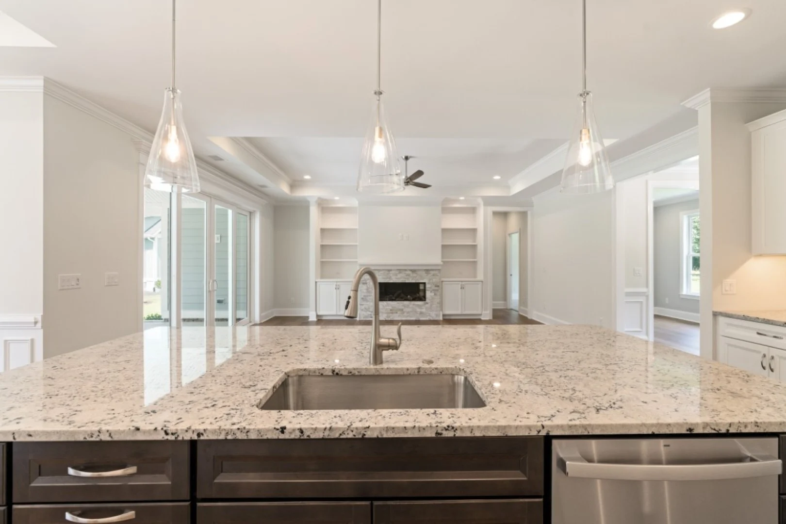 gallery-kitchen (36) Modern kitchen with marble countertop, pendant lights, and stainless steel sink, leading to a bright living room by a home builder.
