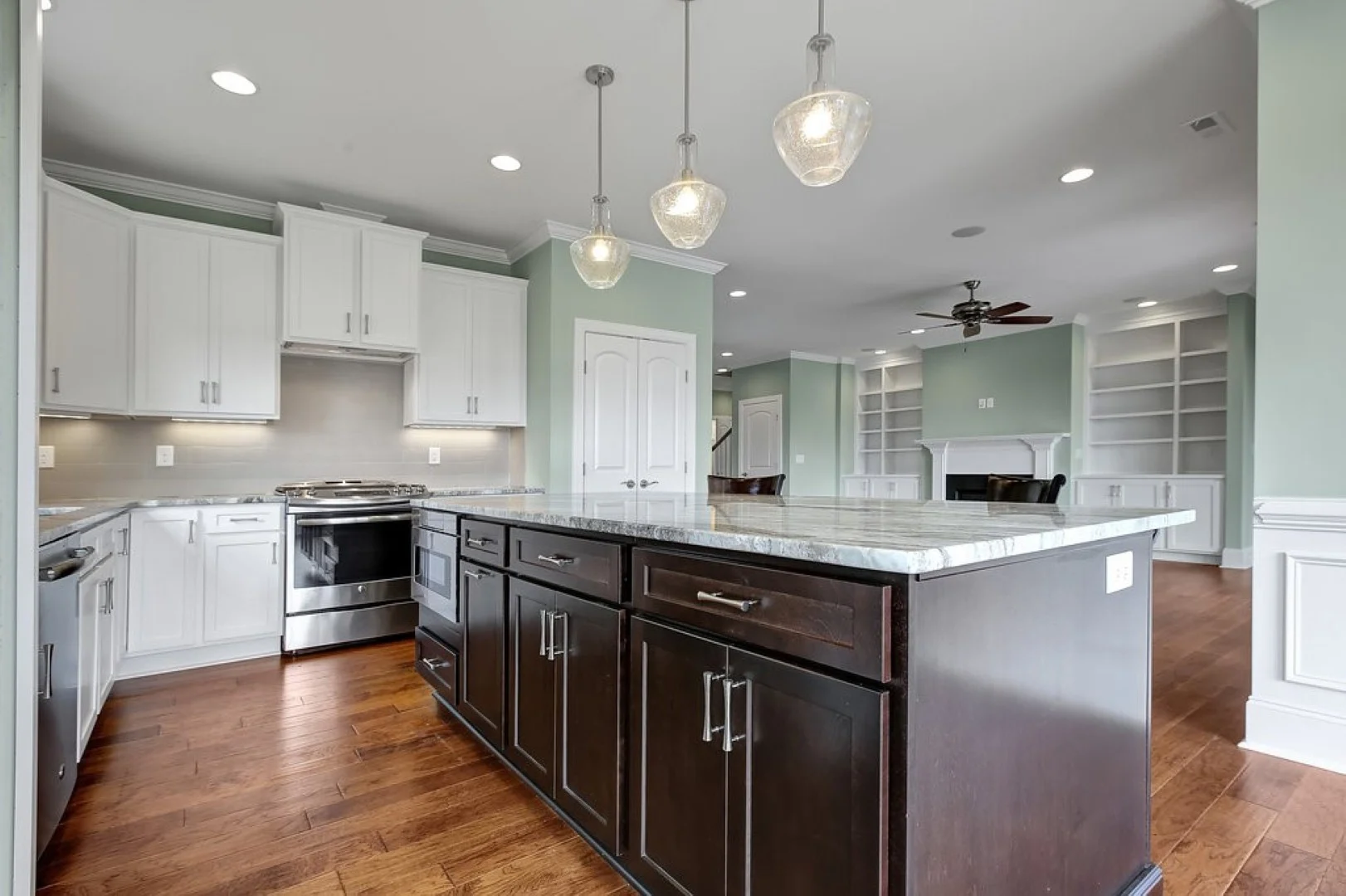gallery-kitchen (47) Spacious kitchen with dark wood island, white cabinets, pendant lights, hardwood flooring, and a glimpse into a living area with built-ins. Ideal for