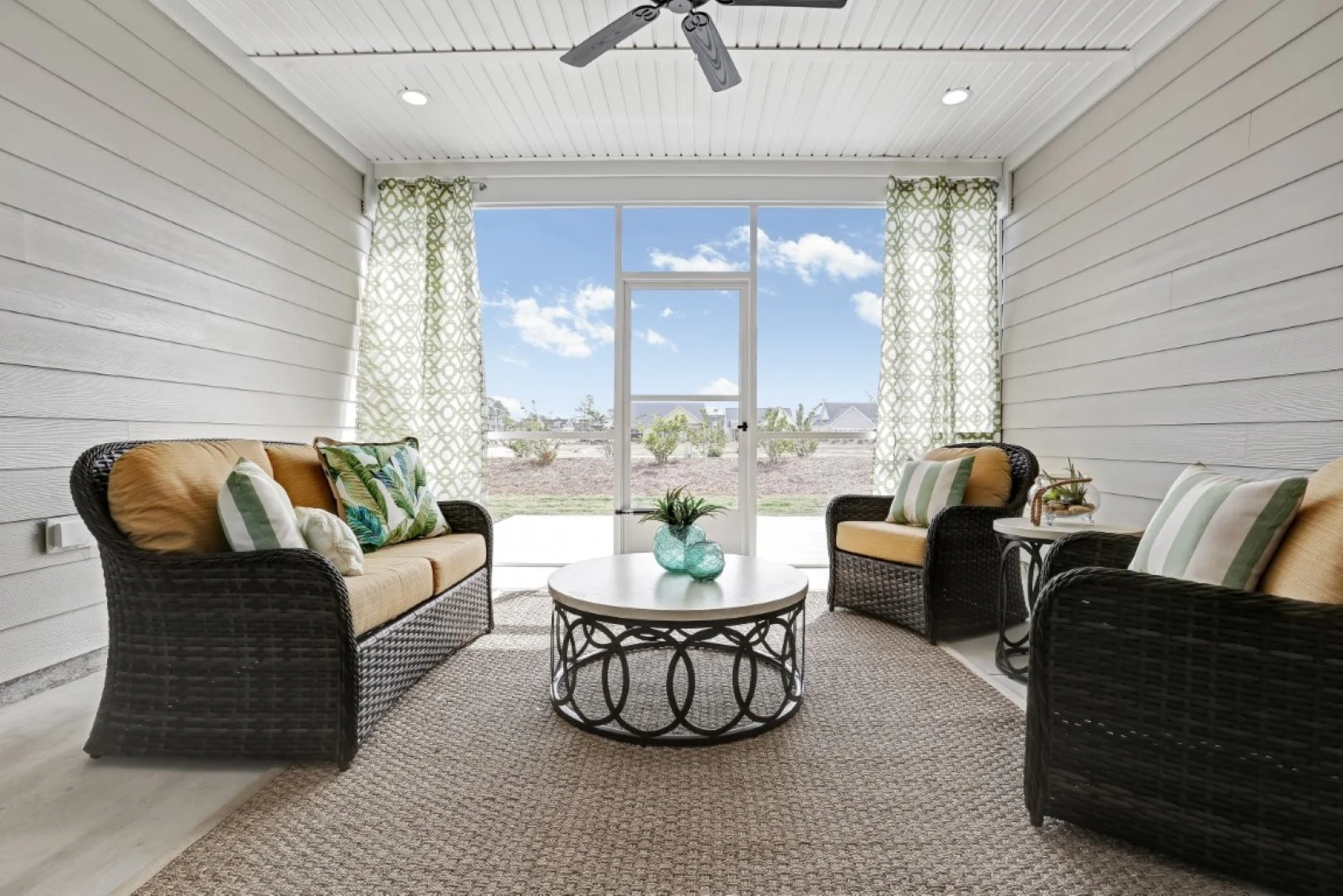 Bright sunroom with wicker furniture, green cushions, and a round table. Large windows overlook a garden. Ideal space by a home builder.