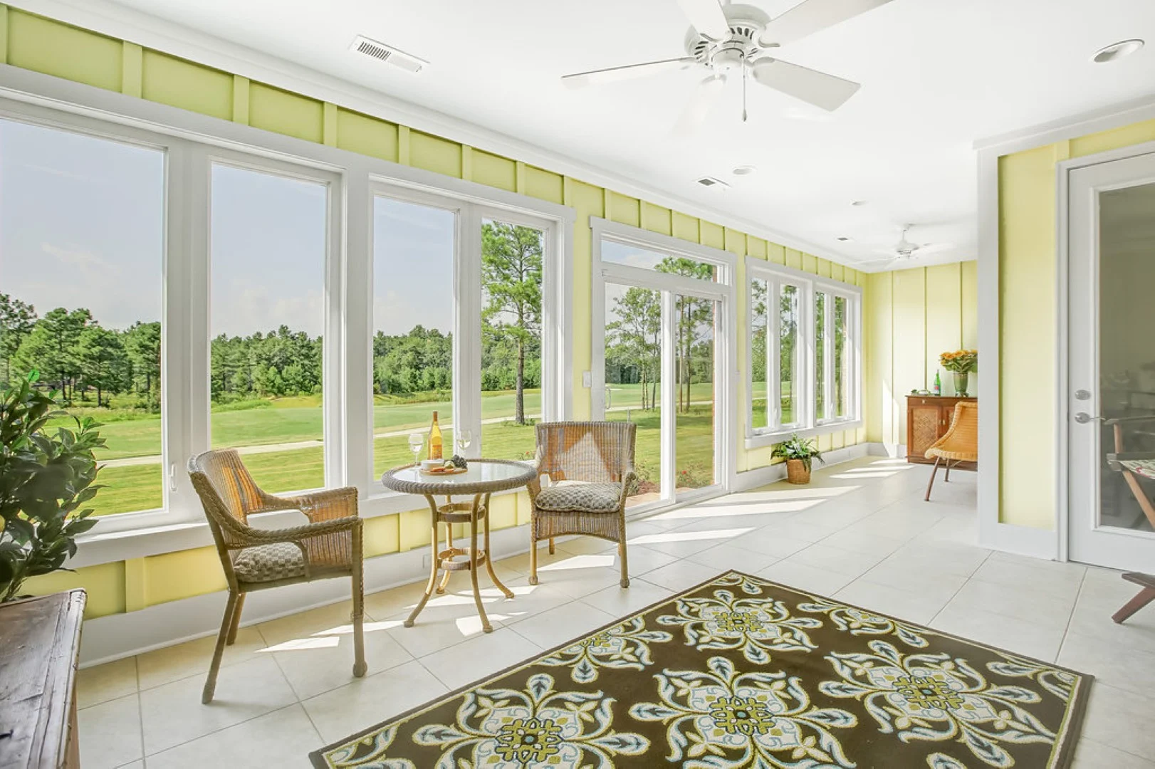 Bright sunroom with large windows overlooking a green landscape, wicker chairs, and a patterned rug, showcasing elegant home builder design.