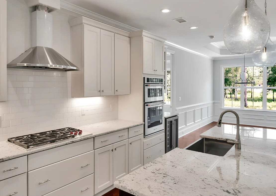 Modern kitchen with white cabinets, marble countertops, and stainless steel appliances, showcasing home builder craftsmanship. Large windows provide n