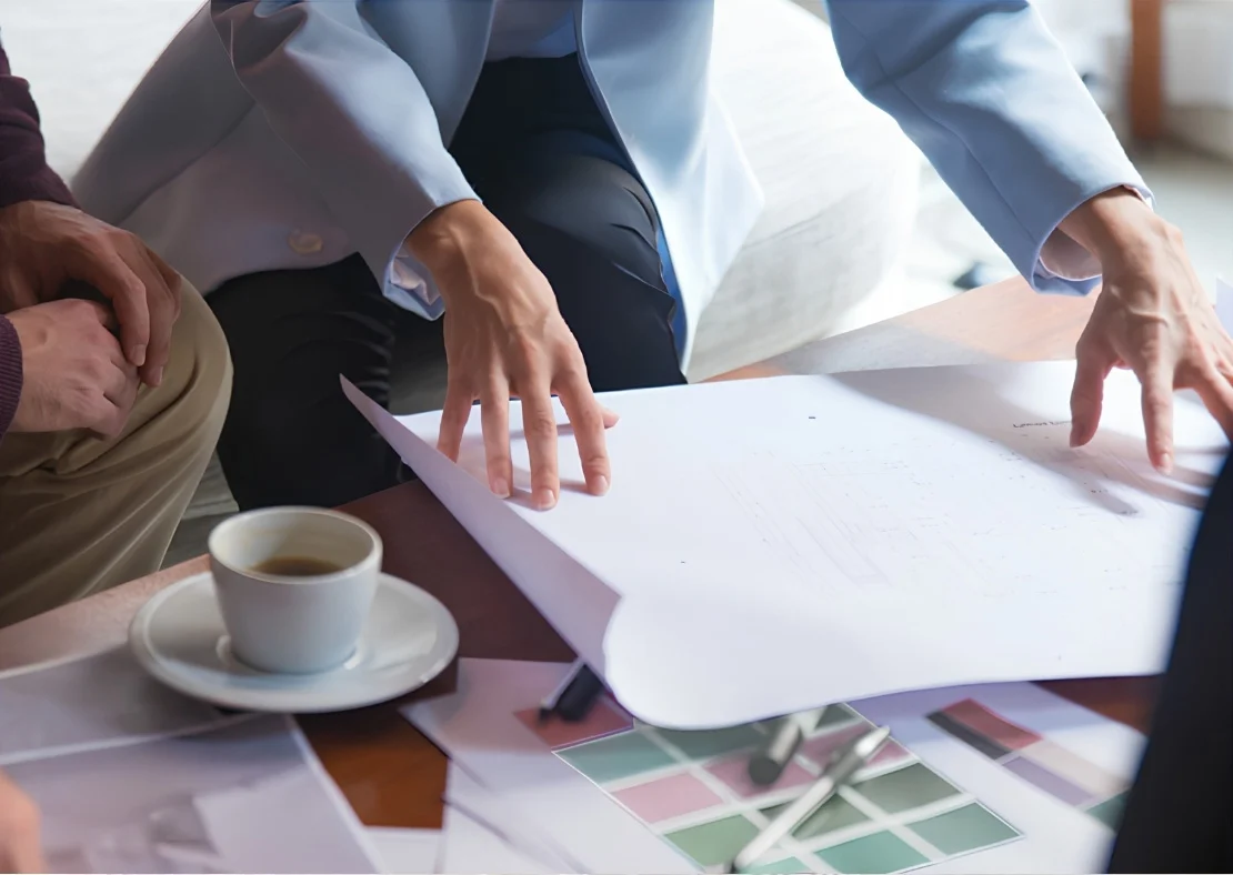 Hands pointing at architectural plans on a table, coffee cup nearby, with color swatches, illustrating a home builder meeting.