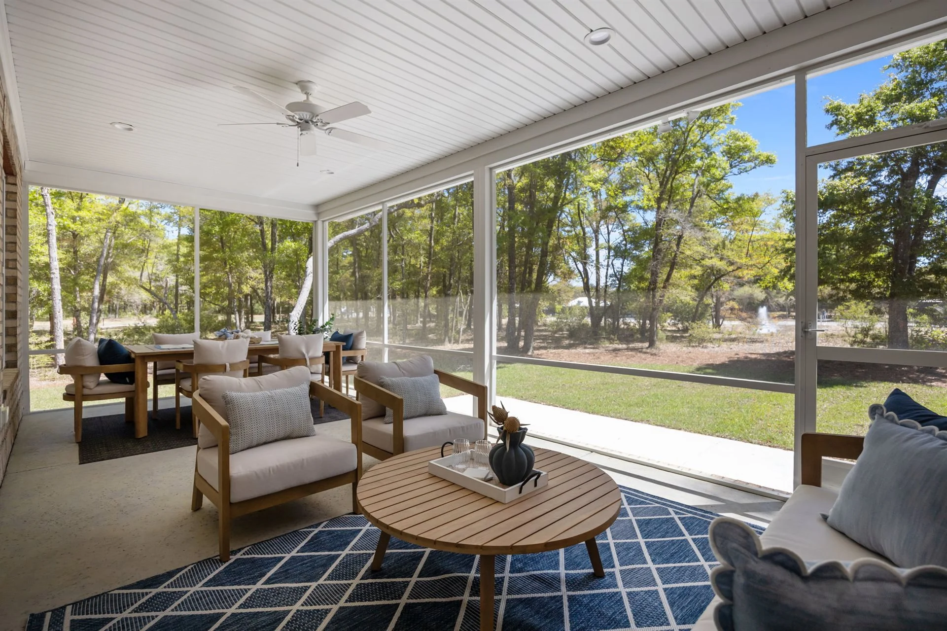 Screened patio with comfortable seating, wooden table, blue rug, and view of lush trees, showcasing modern home builder design.